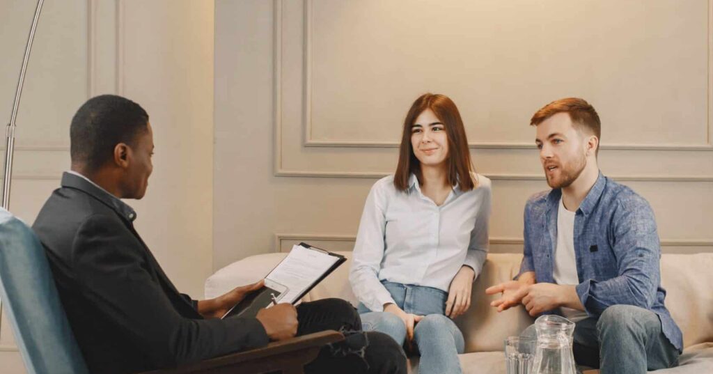 Four adults sit in a modern living room, engaged in a discussion around a coffee table with a curved floor lamp overhead on a shaggy rug.